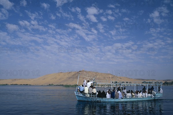 EGYPT, Upper Egypt, Aswan, Qubbet el-Hawa and The Tombs of The Nobles with a loaded  passenger ferry sailing past on the river Nile