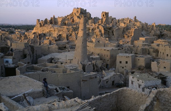 EGYPT, Western Desert, Siwa Oasis, Woman sitting on rooftop with Shali Fortress behind at dusk