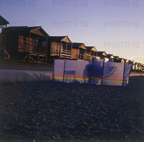 ENGLAND, Kent, Whitstable, Couple silhouetted behind a wind break on the beach in front of wooden beach huts in the  golden evening light
