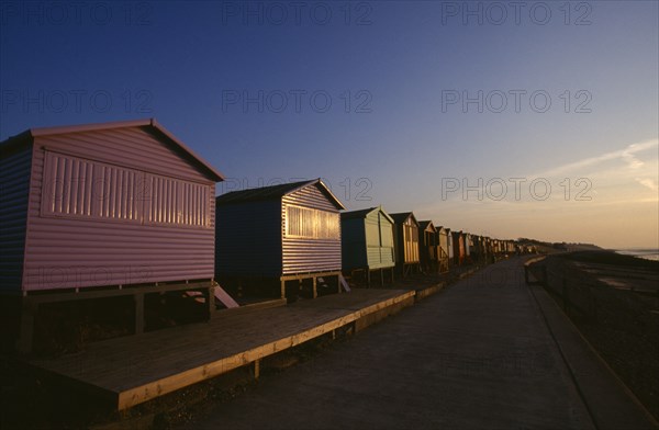 ENGLAND, Kent, Whitstable, View along a row of beach huts painted different colours on Whitstable Beach in golden evening light