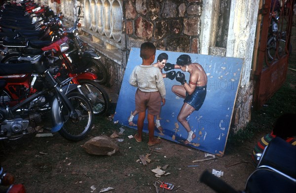 CAMBODIA, Battambang, Small boy looking at painting of boxers propped against the wall in a motorcycle yard.