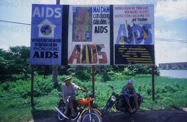 VIETNAM, Hai Duong, Two young men sitting on their motorcycles under a billboard of aids awareness posters.