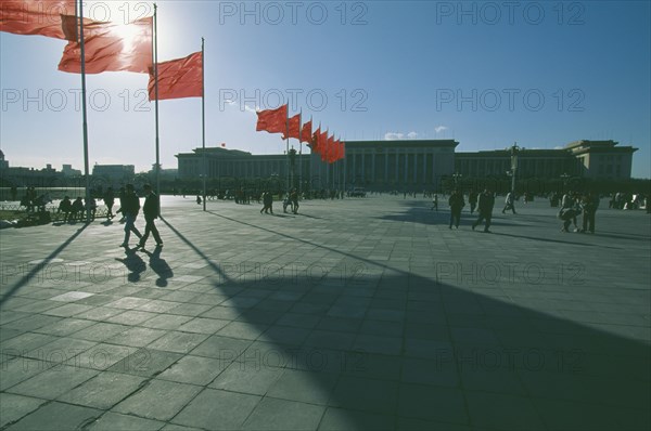 CHINA, Beijing, Tiananmen Square, Great Hall of The People in silhouette with sun behind flying flags and people walking across square with long shadows