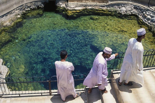 OMAN, Al Rustaq, "Three men in traditional white dishdashas and brimless, circular hats standing beside hot springs."