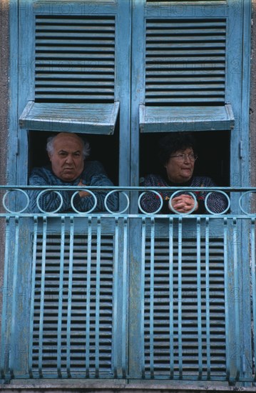 ITALY, Campania, Naples, San Giorgio a Cremano.  Elderly couple looking out from turquoise open wooden window shutters above street.