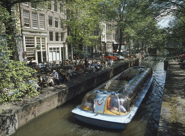 HOLLAND, Noord, Amsterdam, Glass roofed tourist boat passing canal side cafe on the Lelegracht canal