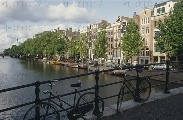 HOLLAND, North, Amsterdam, Bicycles parked beside railings with canal and typical housing behind.