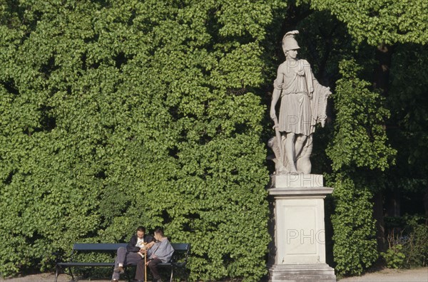 AUSTRIA, Vienna, Schonbrunn Gardens.  Classical statue against hedge with two visitors sitting on bench at one side.