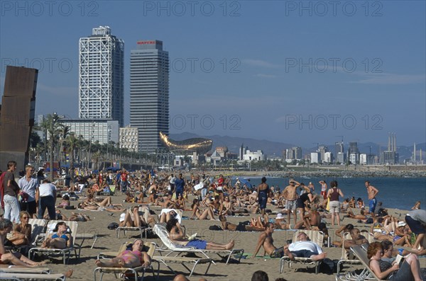 SPAIN, Catalonia, Barcelona, Vila Olimpica area.  Crowded sandy beach with wire mesh monument by Frank Gehry and the Arts Hotel and other skyscraper behind.