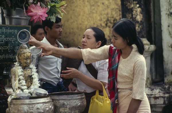 MYANMAR, People, Young women making offerings at Buddhist shrine with small seated Buddha figure garlanded with flowers.