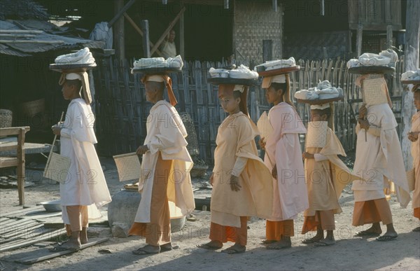 MYANMAR, Mandalay, Buddhist nuns wearing white and orange robes carrying trays of wrapped objects on their heads.