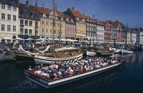 DENMARK, Zealand, Copenhagen, Nyhavn Canal. Cruise boat passing sailing boats moored on the quayside beside brightly painted traditional houses
