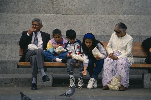 ENGLAND, London, Trafalgar Square, Asian family sitting together on a bench eating with girl leaning forward to feed a pigeon