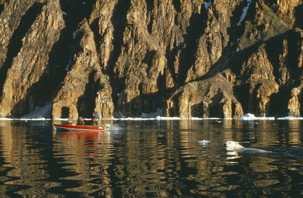 CANADA, Baffin Island, Eskimos in motor boat and Polar Bear fishing in water against a backdrop of cliffs