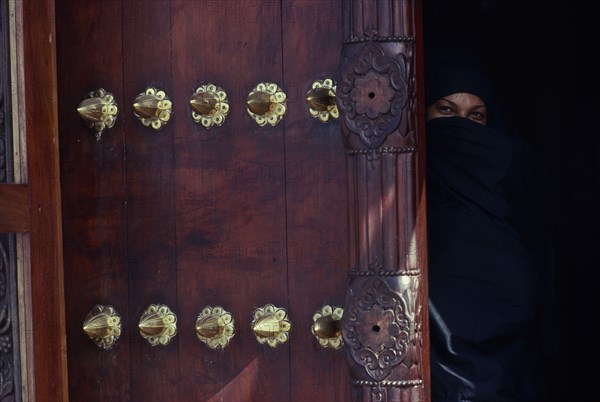 ZANZIBAR, People, Woman in full chadoor looks out from behind ornate carved and studded door.