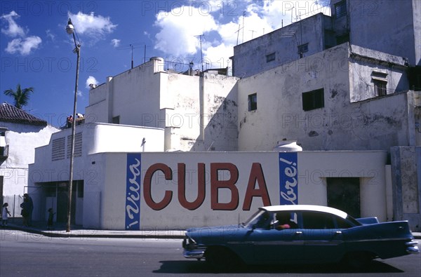 CUBA, Havana, Street scene with passing classic car and the words Viva Cuba painted on the wall opposite