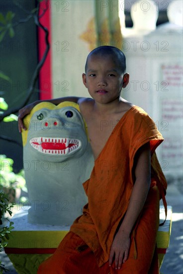 LAOS, Savannakhet, Wat Sainyamungkhun. Young monk sitting beside colourful statue at the Temple