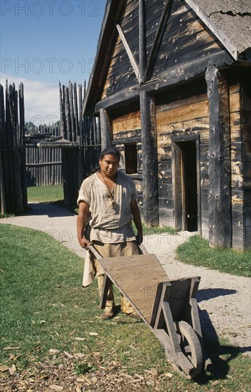 CANADA, Ontario, Indigenous people, Sainte Marie among the Hurons near Midland. Indian man pushing a wheel barrel near wooden building.