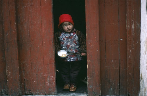 CHINA, Guangxi, Yangshuo, Child holding bowl of rice and chopsticks looking out from narrow doorway.