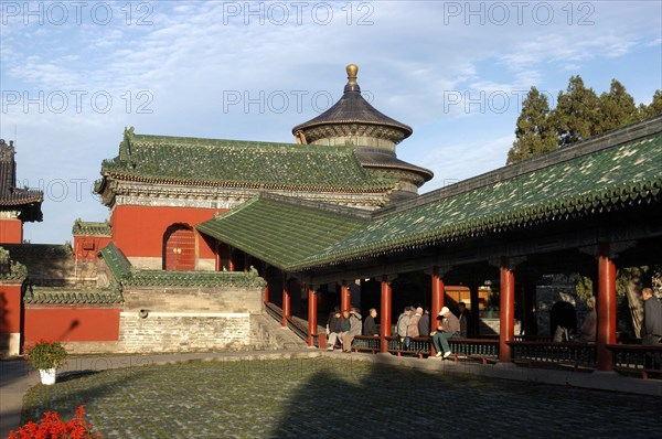 CHINA, Beijing, Tiantan Park, aka The Temple of Heaven. View of the red and green rooved architecture within the complex with people sitting on a fence