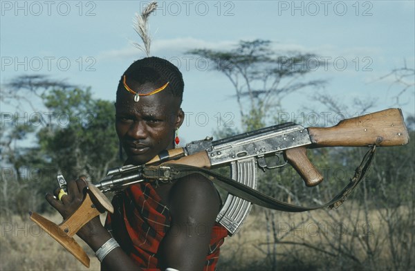 UGANDA,   , Karamoja, "Portrait of Karamojong warrior with ostrich feather in hair holding gun and stool, always carried and used as both seat and head rest. "