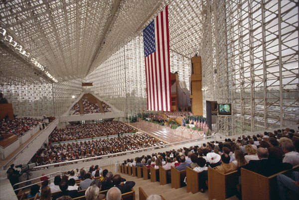 USA, California, Los Angeles, Crystal Cathedral Memorial Service. Congregation in pews under glass structure beneath flag of Stars And Stripes and a large TV