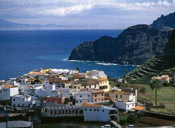 SPAIN, Canary Islands, La Gomera, "Agulo.  White yellow and orange painted houses with tiled rooftops, terraced hillside volcanic cliffs and sea beyond."