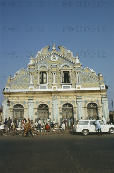 NIGERIA, Lagos, Street scene with roadside vendors ad passers by in front of mosque with pale blue and cream exterior decorated with Islamic relief carving.