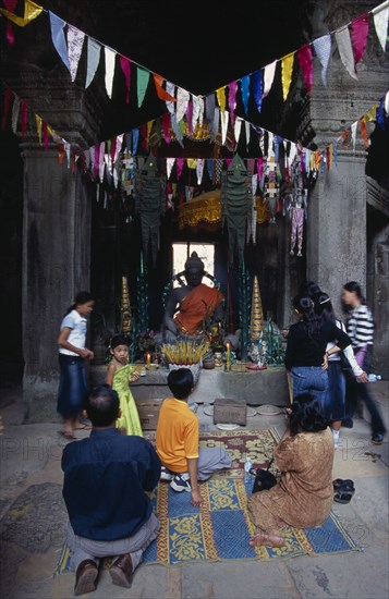 CAMBODIA, Siem Reap Province, Angkor, Banteay Kdei.  Cambodian visitors lighting incense at shrine with seated Buddha figure hung with coloured flags for Chinese New Year.