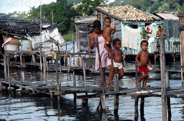 BRAZIL, Bahia, Salvador da Bahia, Young woman with three children crossing wooden bridge over water polluted with sewage in shanty town.  Houses raised on stilts above water behind.