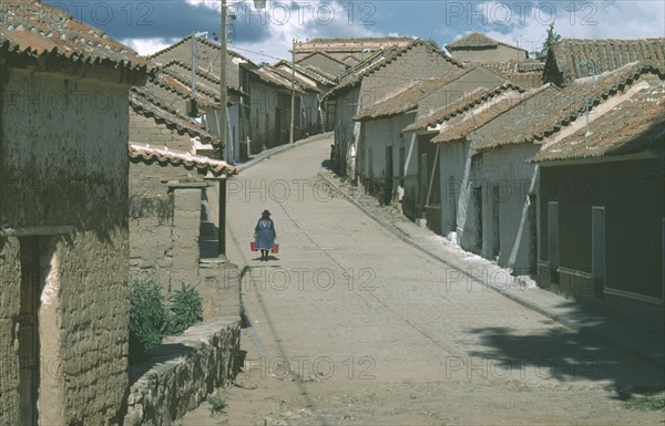 BOLIVIA, Chuquisaca, Tarabuco, Town in the Altiplano or Bolivian Plateau.  Woman carrying two buckets walking along narrow paved street between houses with overhanging tiled rooftops.