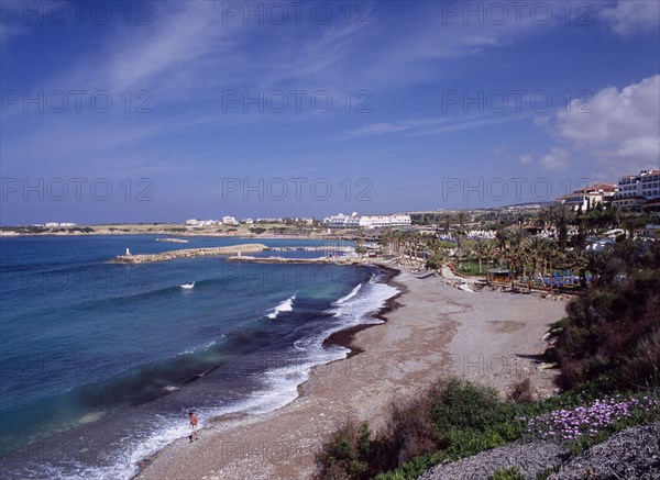 CYPRUS, Coral Bay, Man walking along shore of quiet beach with small harbour behind lined by palms and promenade and overlooked by resort buildings.