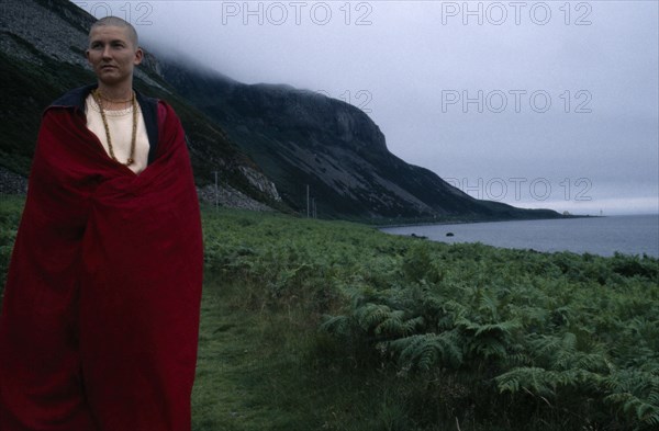 SCOTLAND, Clyde, Tibetan Nun draped in a red robe standing next to coastline by green landscape and mountains at a Retreat on Holy Island.