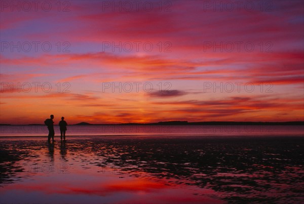IRELAND, Sligo, Rosses Point, Two figures silhouetted on the beach watching dramatic deep pink and orange sunset reflected on wet sand.