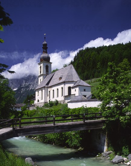 GERMANY, Bavaria, Ramsau, View over river and wooden bridge towards Ramsau Church and wooded hillside beyond.