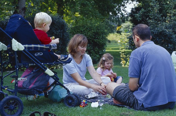 ENGLAND, West Sussex, Chichester, The Bishops Palace Gardens.  Young family having picnic.  Parents with daughter aged three and son aged one and a half in push chair.