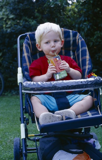 ENGLAND, West Sussex, Chichester, The Bishops Palace Gardens.  Boy aged one and a half drinking from carton of low sugar blackcurrant drink while sitting in pushchair during picnic lunch.