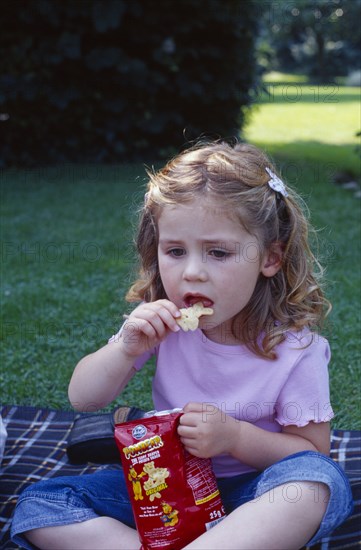 ENGLAND, West Sussex, Chichester, The Bishops Palace Gardens. Girl aged three eating potato crisp snack during picnic lunch.