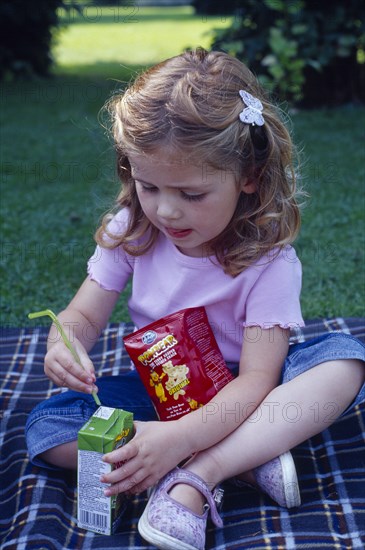 ENGLAND, West Sussex, Chichester, The Bishops Palace Gardens.  Girl aged three putting straw into carton of low sugar blackcurrant juice drink during picnic lunch.