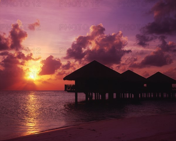 MALDIVES, Baros Island, View of silhouetted beach huts on stilts over water against a dramatic pink setting sky