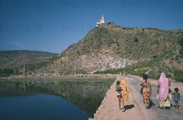 INDIA, Rajasthan, General, Women and children walking beside water reservoir tank twenty miles north of Udaipur.  Hindu shrine on hill top beyond.