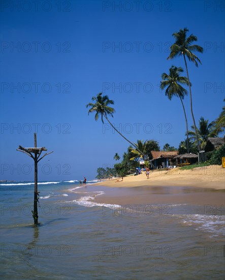 SRI LANKA, Hikkaduwa, View along sandy beach with overhanging palms toward beach front restaurant and fishermans perch in the foreground