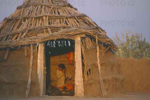 INDIA, Rajasthan, Thar Desert, Bhikodai.  Early morning light on thatched mud hut with woman plaiting her hair framed in doorway.