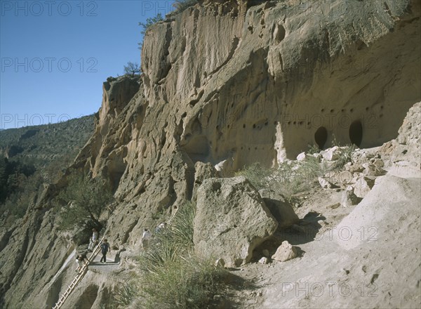 USA, New Mexico, Bandelier National Monument. , Visitors climbing ladder up a cliff towards the caves.