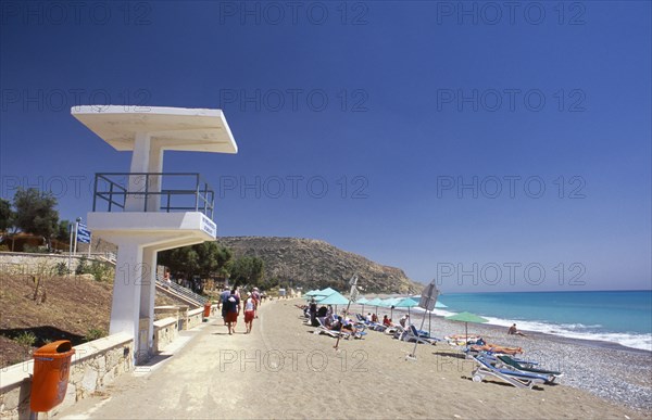 CYPRUS, Pissouri Bay, Sunbathers on narrow sand and pebble beach with people walking along promenade at side and life guards tower or look out point in the foreground.
