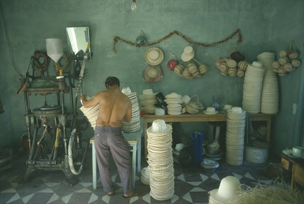 COLOMBIA, Narino Department, Sandona, Panama hat industry.  Male worker operating machinery with hats stacked on floor and table beside him.