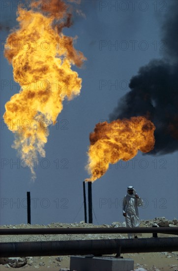 SAUDI ARABIA, Industry, Flaming gas on oil field with man standing behind pipe in foreground.