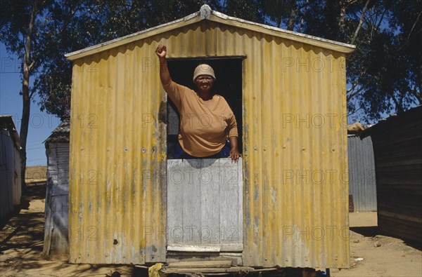 SOUTH AFRICA, Western Cape, Malmsbury, Woman standing with her fist in air looking out of a yellow corrugated hut a resident of Malmsbury council Hostel.