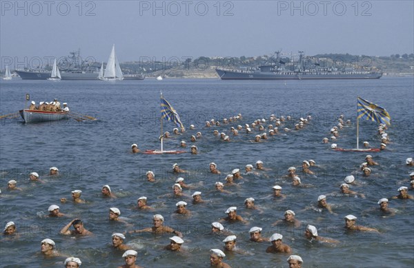 RUSSIA, Sevastopol, Navy Day. Sailors swimming in the sea in formation with battleships behind.