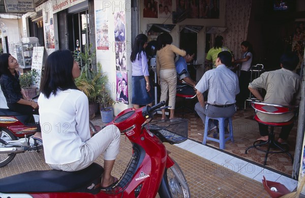CAMBODIA, Siem Reap, Busy hairderssers shop with two women sitting on mopeds watching a television in the back of the shop